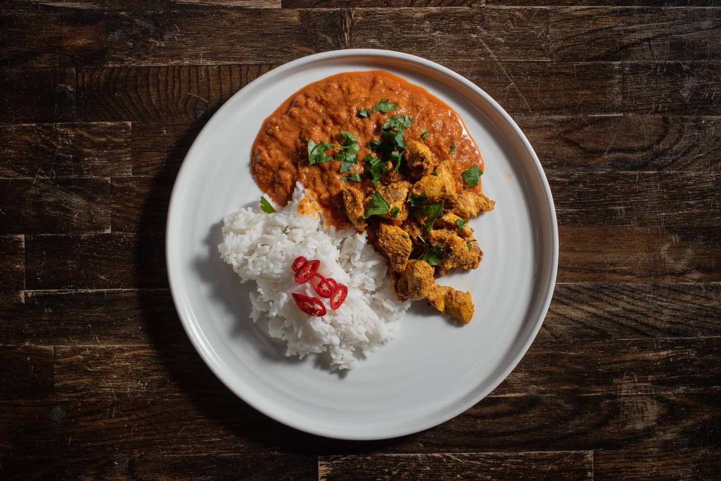 Kawałki kurczaka w przyprawach z natką, pomarańczowy sos i ryż z krążkami chili na białym talerzu