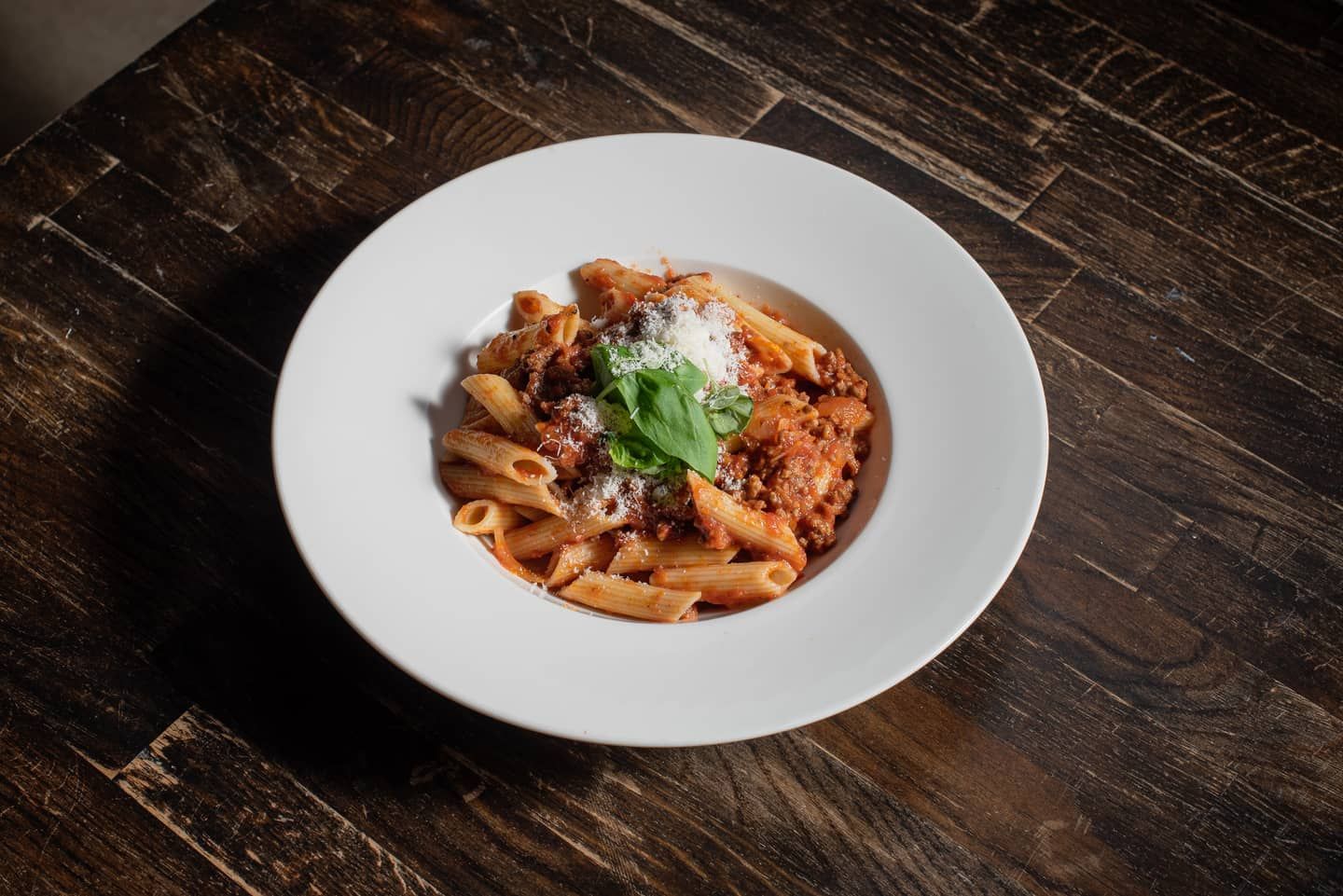 Głęboki talerz z penne w sosie mięsnym, posypane parmezanem i udekorowane bazylią, na ciemnym drewnie